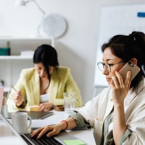 Two women multitasking in a contemporary office setting, balancing phone calls and laptops.